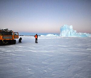 Two expeditioners beside a tracked vehicle looking right towards an iceberg encased in sea ice