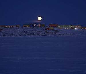 Moon rising over station with buildings in profile from sea ice