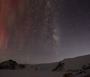 Nightsky visible with red and green aurora hanging suspended above the outline of a plateau