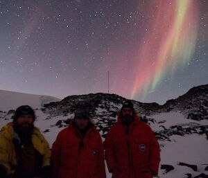 Three expeditioners pose in front of a hut with an aurora in the sky behind them