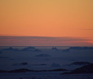 Orange sky above blue of the sea ice with icebergs silhouetted