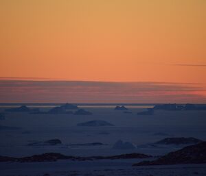 Orange sky above blue of the sea ice with icebergs silhouetted