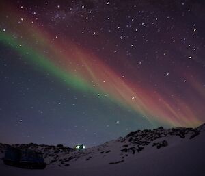 Green and red auroras above a rocky hill with a green field hut illuminated at night and unlit tracked vehicle in foreground