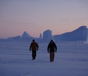 Two expeditioners in foreground walking towards camera with iceberg in background