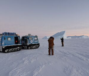 Tracked vehicle in foreground with two expeditioners taking photo of iceberg in background