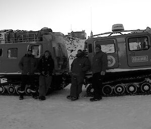 Four expeditioners standing in front of tracked vehicle with hut in background