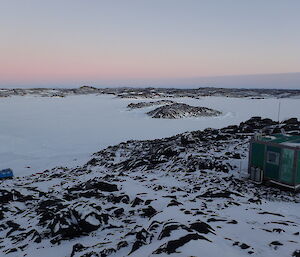 Rocky terrain capped with snow, green field hut to the right in foreground. Blue tracked vehicle in left background