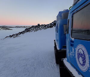 Blue Häggulands overland vehicle parked at right of photo foreground with sea ice and rocks in background