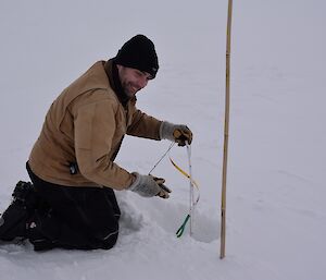 Expeditioner kneeling beside a drill hole measuring the thickness of ice with a tape measure