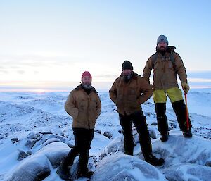 Three expeditioners standing in rocky terrain covered in snow facing camera smiling