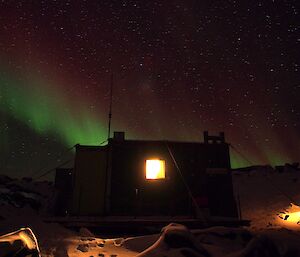 Hut with light streaming from windows outlined against a night sky dotted with stars and the green and red glow of an aurora