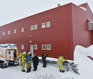 Group of expeditioners in fire fighting gear next to a tracked vehicle beside a large red accommodation building