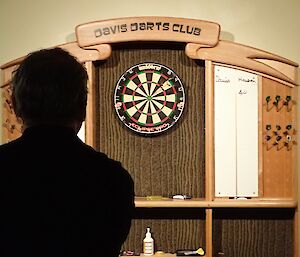 An expeditioner in silhouette about to launch a dart at the board