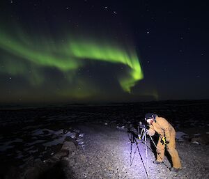 Expeditioner crouching next to a camera on a tripod on a road at night with a green aurora above him