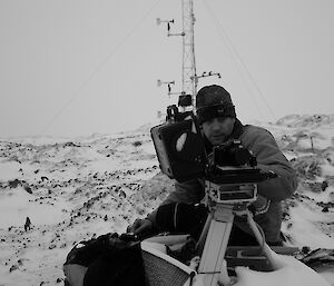 Expeditioner sitting beside an automated camera with an antenna in background on rocky snow covered terrain