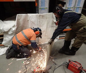 One expeditioner kneeling on the floor whilst another holds a shielding board whilse working on a railing with a grinder
