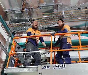 Two expeditioners looking down from a scissor lift as they work on piping