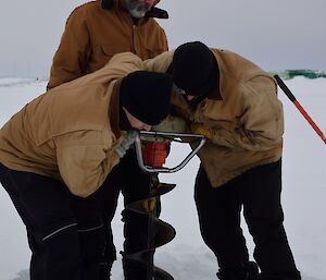 Three expeditioners leaning over a large ice drill