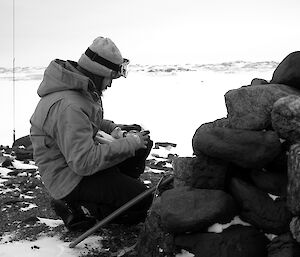 Expeditioner crouches in profile with the base of the cross in foreground as he reads messages left within a capsule