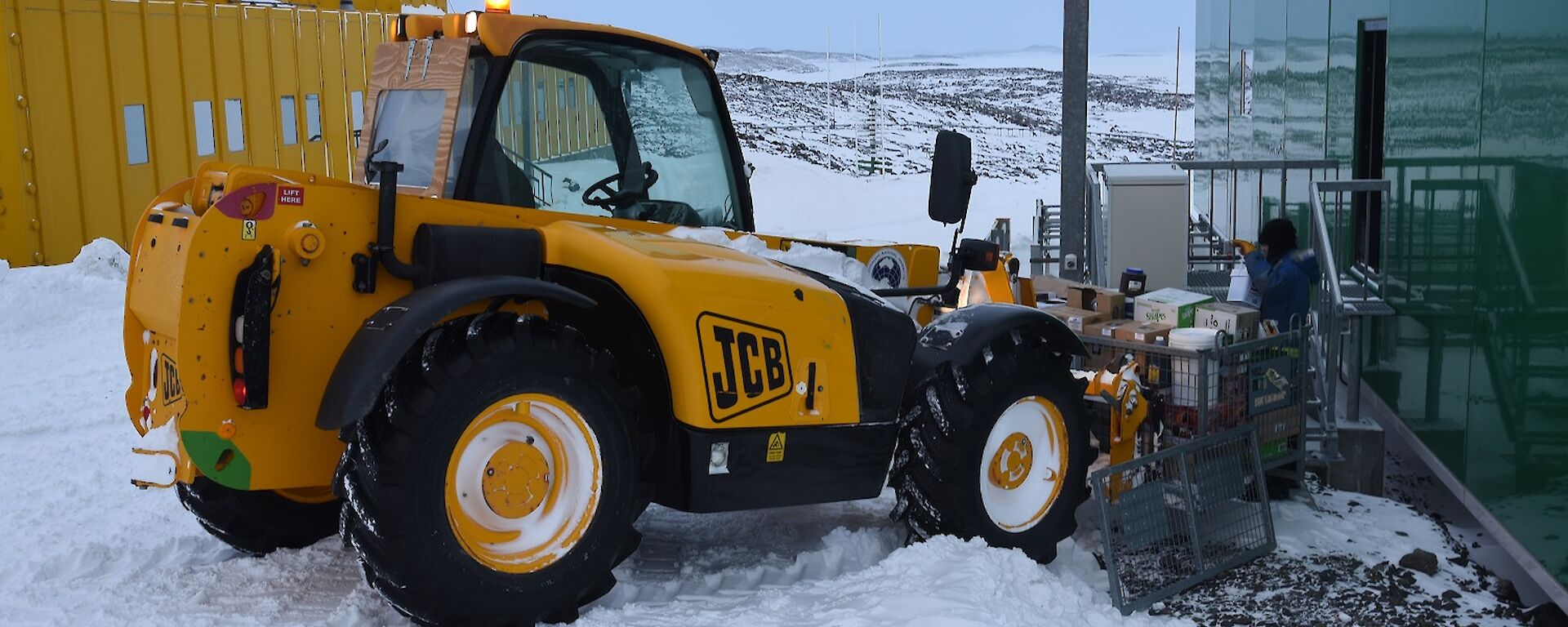 Expeditioner standing on steps of a green building with a JCB digger raising a cage pallet of food supplies.