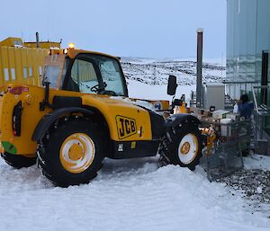 Expeditioner standing on steps of a green building with a JCB digger raising a cage pallet of food supplies.