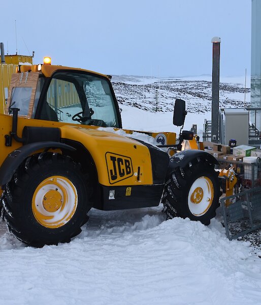 Expeditioner standing on steps of a green building with a JCB digger raising a cage pallet of food supplies.