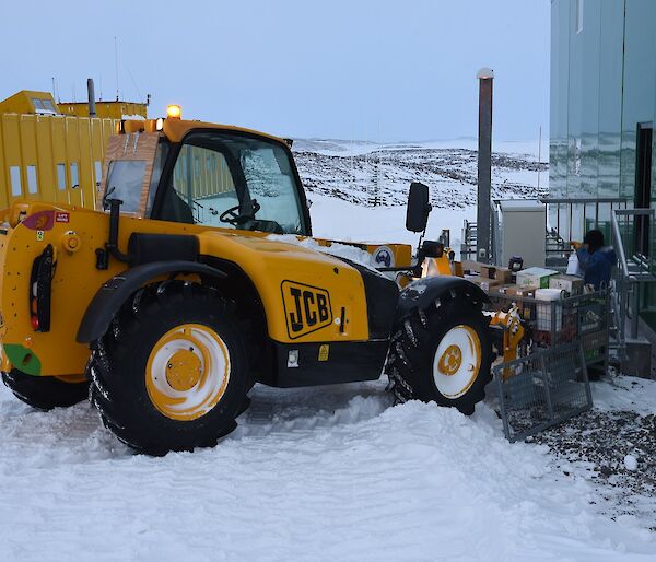 Expeditioner standing on steps of a green building with a JCB digger raising a cage pallet of food supplies.