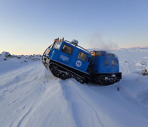Blue Hägglunds tracked vehicle rising up over a snow bank with rocky hills in the background.