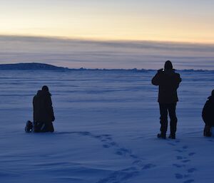 Expeditioners stand and kneel in the foreground as an emperor penguin stands in the background.