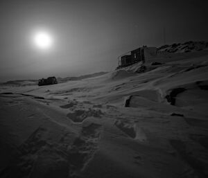 A night shot with the moon illuminating a hut and Hägglunds tracked vehicle amidst snowfall.