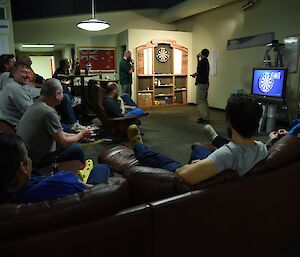 Expeditioners seated at the bar and on couches, facing a televised darts competition.