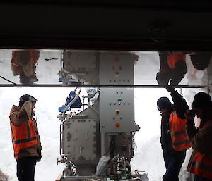 Three expeditioners silhouetted against the bright entrance of a basement, exchanging puzzles glances as to how to manouver a piece of equipment inside.