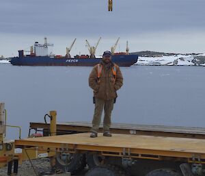 Expeditioner standing on a flat top truck with ship and harbour in background.