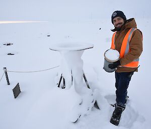 Expeditioner standing with a metal drum full of snow beside a measuring gauge.