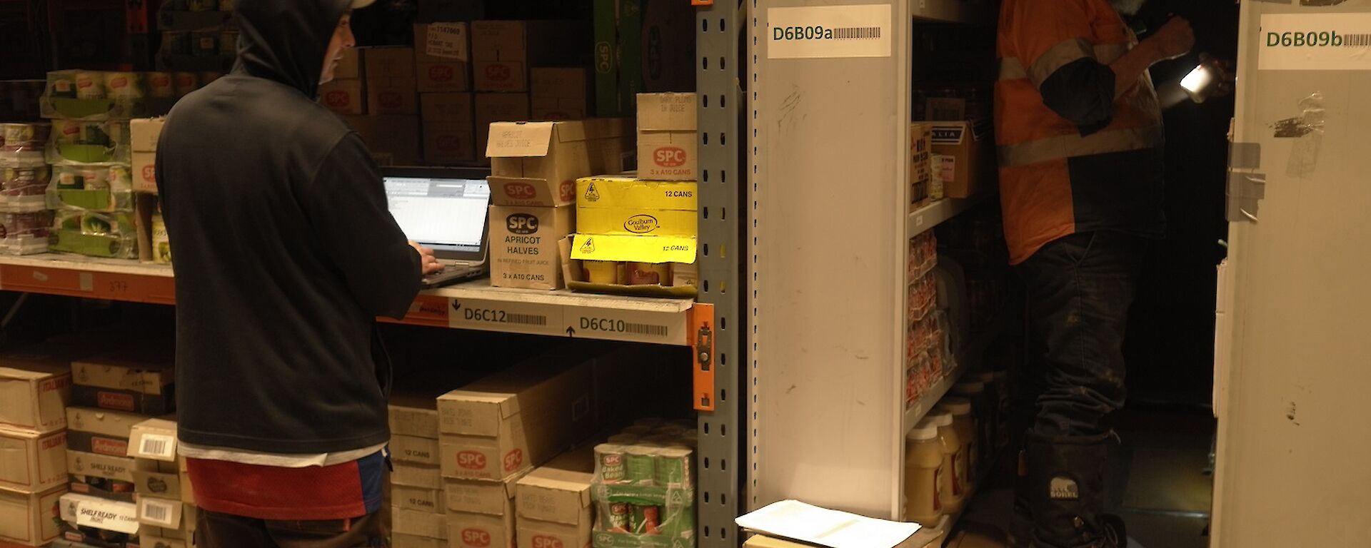 Expeditioner standing beside shelving with a laptop and another expeditioner in an aisle holding a flashlight.