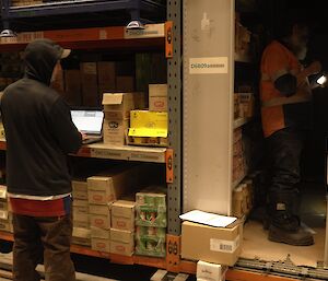 Expeditioner standing beside shelving with a laptop and another expeditioner in an aisle holding a flashlight.