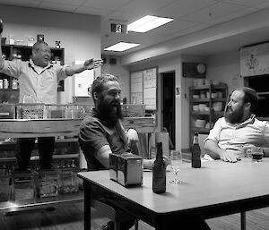 Expeditioner holding a bottle, gesturing behind a bar as two expeditioners look on smiling.