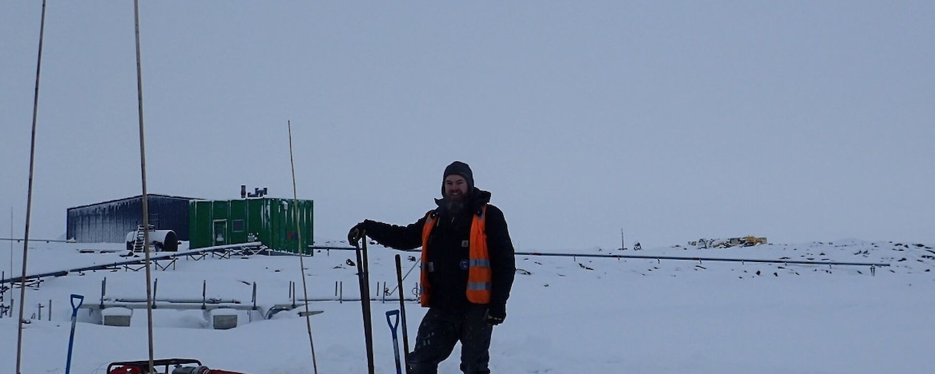 Expeditioner standing shovel in hand next to a water pump and generator with a green building in the background