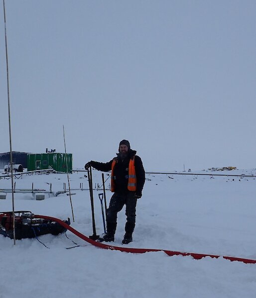 Expeditioner standing shovel in hand next to a water pump and generator with a green building in the background