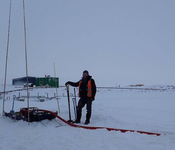 Expeditioner standing shovel in hand next to a water pump and generator with a green building in the background