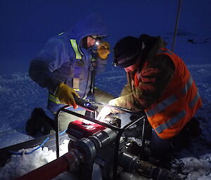 Expeditioner crouches beside a pump holding a light whilst another expeditioner undertakes maintainance on the pump