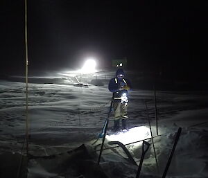 Expeditioner standing over a hole in the snow with a tracked vehicle light in the background