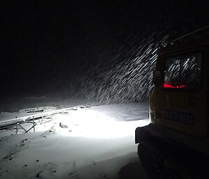Headlights of a tracked vehicle illuminate falling snow and site services piping