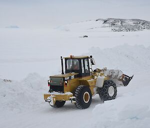 Expeditioner operating a mechanical loader pushing snow in foreground with the sea ice and islands in background