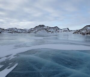 A frozen blue ice lake with snow covered hills in the background