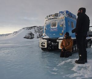 Two expeditioners facing away from camera standing on sea ice with a tracked vehicle in background