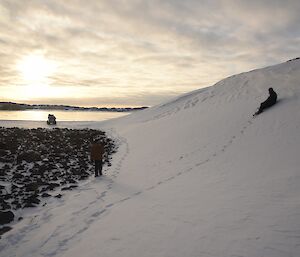 Expeditioner sitting on a snow scour with others walking below towards a frozen lake and tracked vehicle