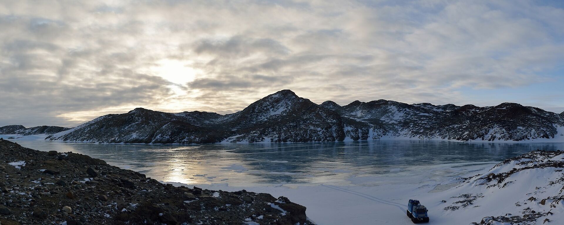 Panoramic photo of a frozen lake and rocky hills with a tracked vehicle in bottom right of frame