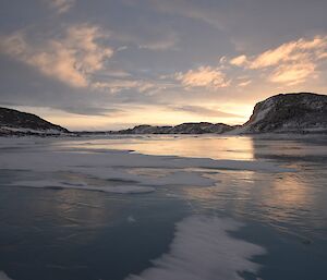 Frozen fjord with rocky hills in background backlit by light from setting sun