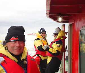 Expeditioner standing on a motor launch foreground with two expeditioners and a ship in the background